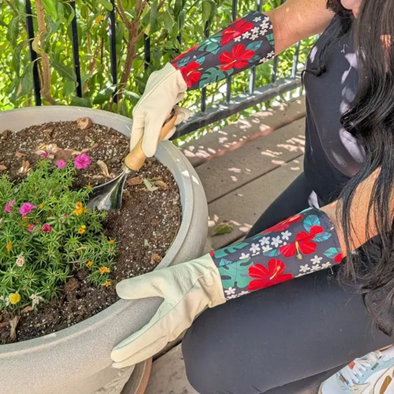 Leder-Gartenhandschuhe mit langen, geblümten Stulpen | Hibiskusblüten | beim Arbeiten mit einer gezahnten Handschaufel in einem großen Pflanztopf